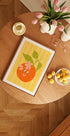 a wooden table with a framed picture of an orange, a bowl of lemons, a vase of pink tulips, and a candle on it.
